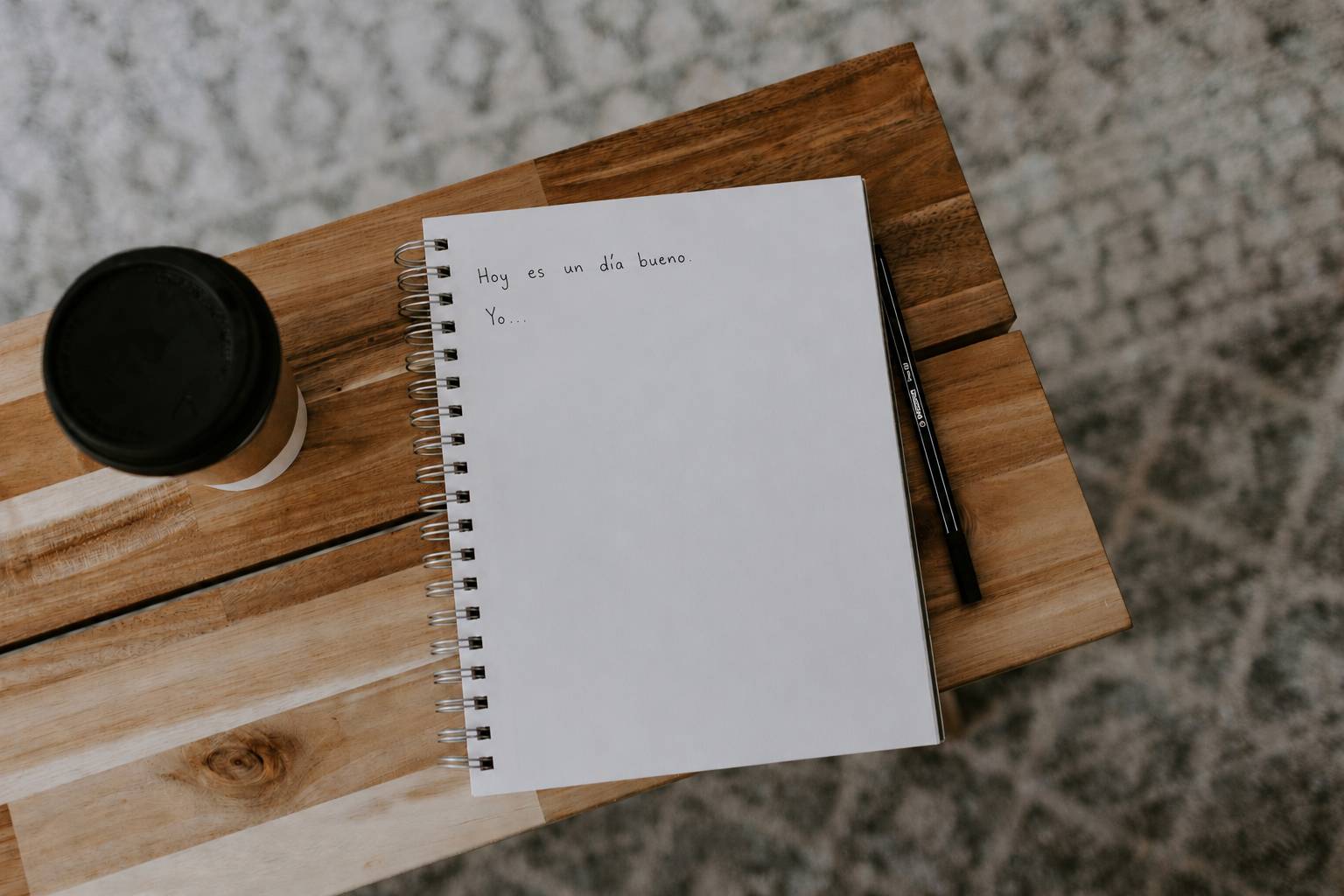 A handwritten Spanish journal entry, 'Hoy es un día bueno. Yo...', open on a wooden table next to a coffee and a pencil