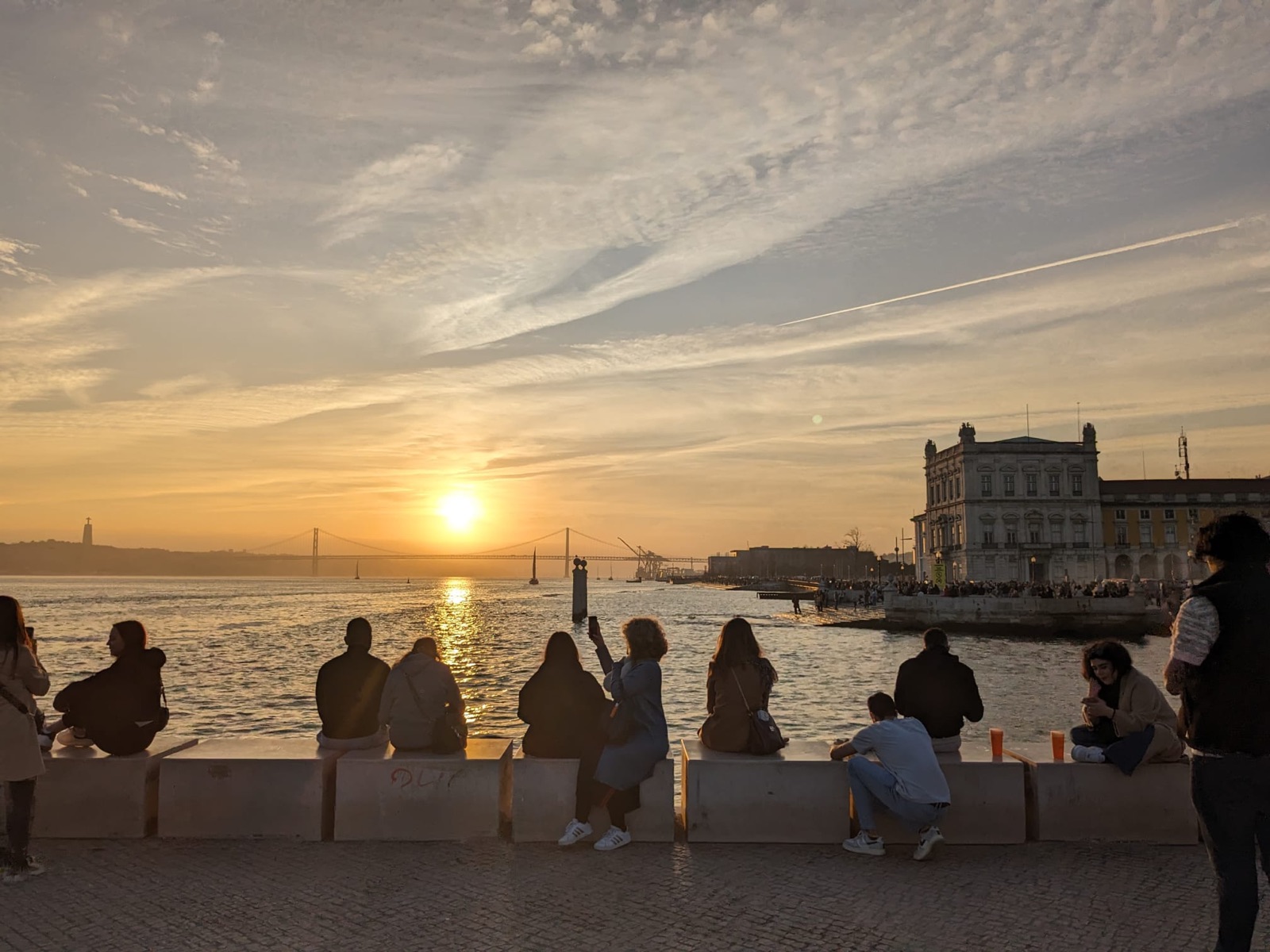 People watching the sunset over the Tagus river at Praça do Comércio in Lisbon, with the 25 de Abril bridge in the distance