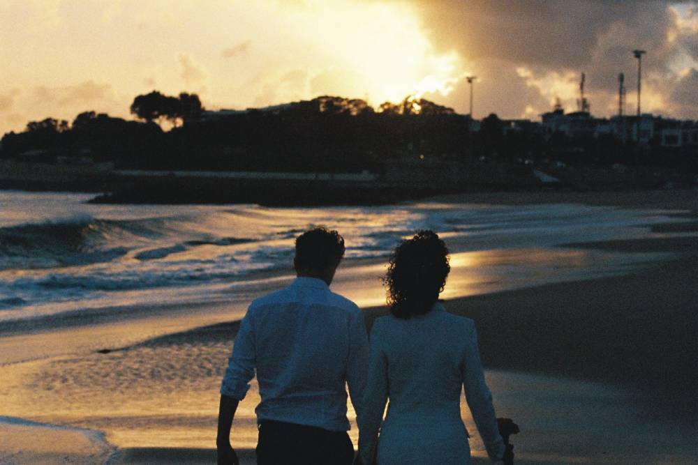 A couple walking along a beach near Lisbon, Portugal at sunset in wedding clothes, seen from behind